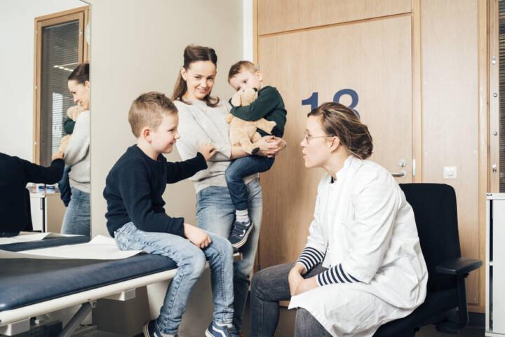 lääkäri tutkii lasta ja äiti seisoo taustalla toinen lapsi sylissään