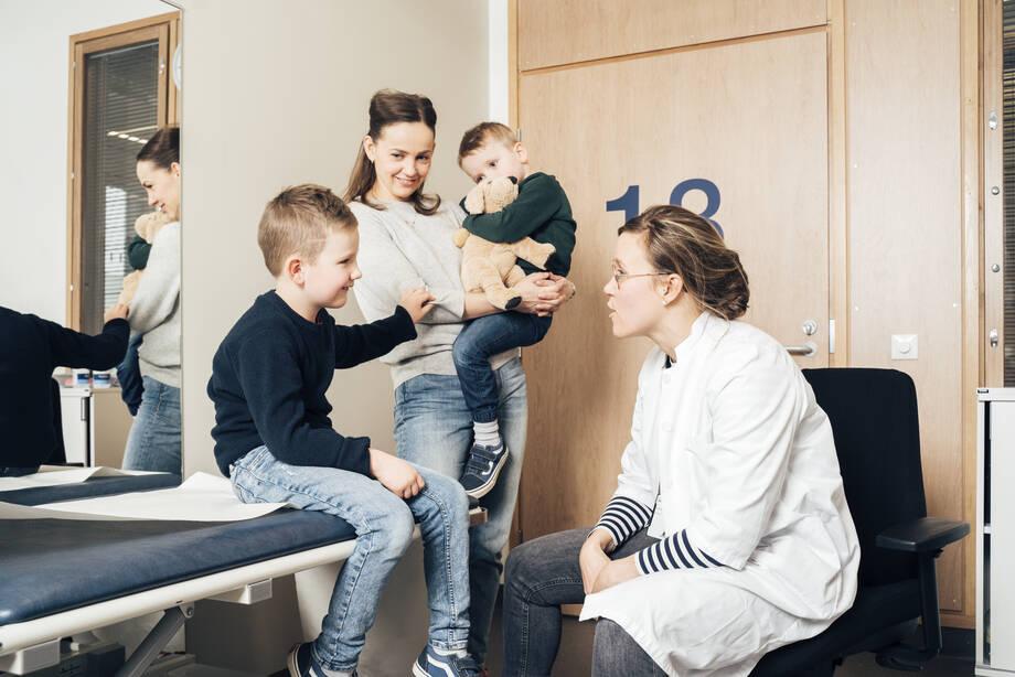 lääkäri tutkii lasta ja äiti seisoo taustalla toinen lapsi sylissään